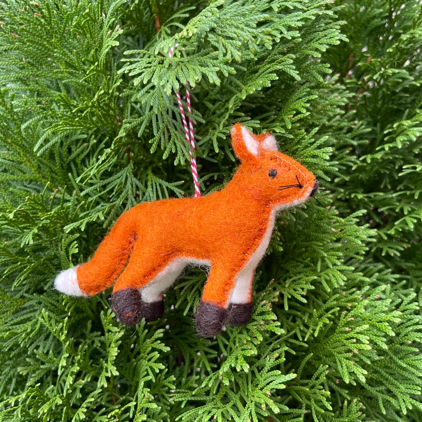 A felted orange fox Christmas ornament with white and dark brown accents, hanging from a red and white striped string amidst green pine needles.