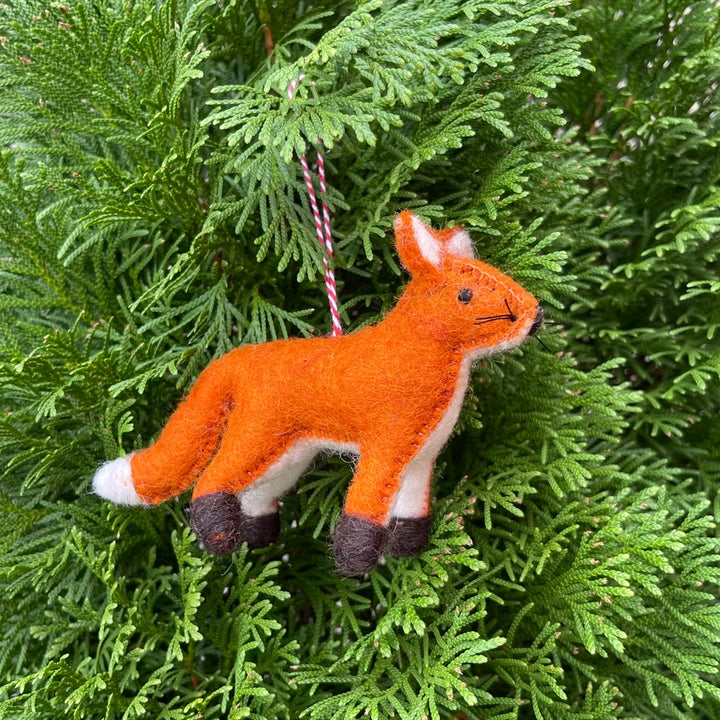 A felted orange fox Christmas ornament with white and dark brown accents, hanging from a red and white striped string amidst green pine needles.