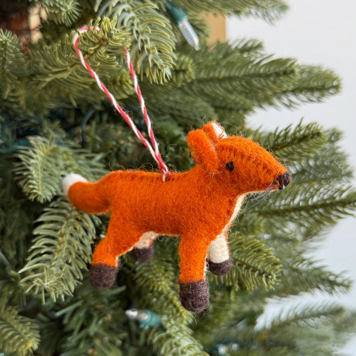 A small felted orange fox Christmas ornament with white and dark brown accents, hanging from a red-and-white striped string on a green evergreen tree.