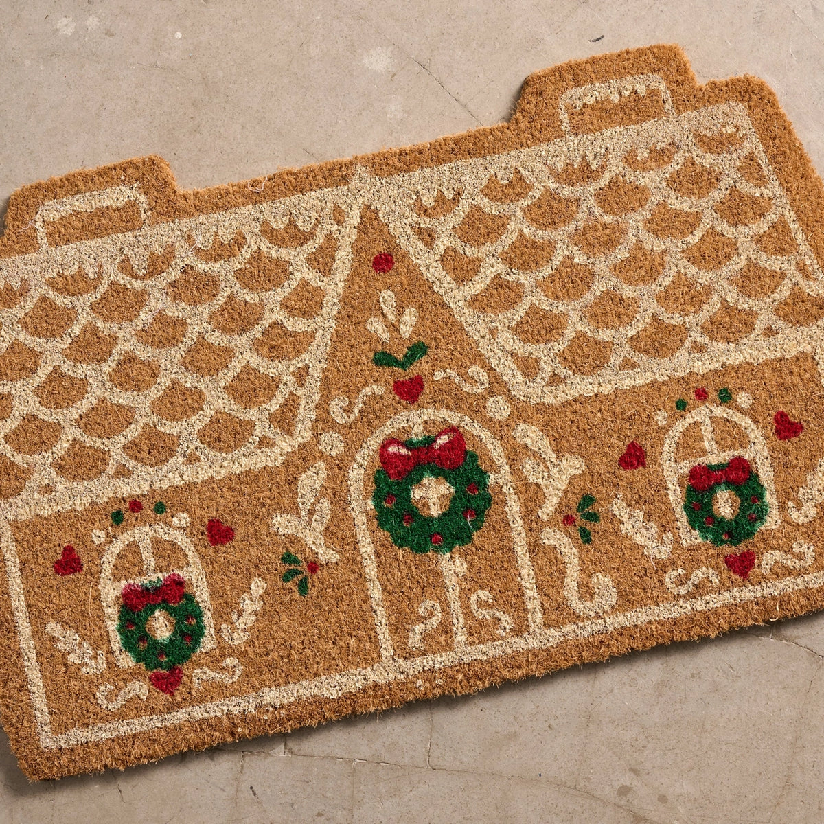 A gingerbread house shaped doormat in natural coir with cream outlines and festive green and red wreath details, photographed on a light concrete floor.