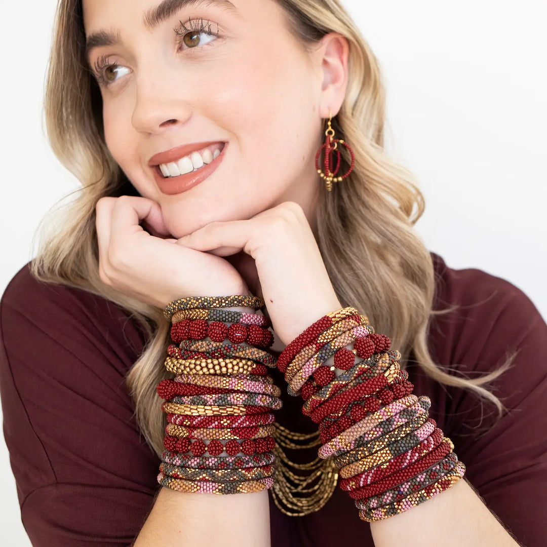 A woman wearing multiple beaded roll on bracelets on both wrists and red hoop earrings, posed against a clean white background.