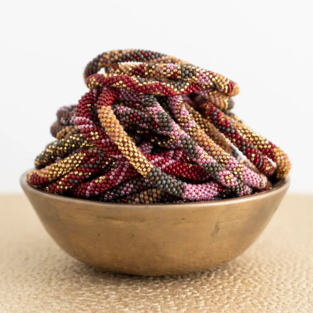 A pile of braided beaded roll on bracelets in mixed earthy colors, including burgundy, gold, brown, and pink, displayed in a small metallic bowl on a textured beige surface against a white background.