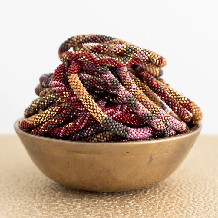 A pile of braided beaded roll on bracelets in mixed earthy colors, including burgundy, gold, brown, and pink, displayed in a small metallic bowl on a textured beige surface against a white background.