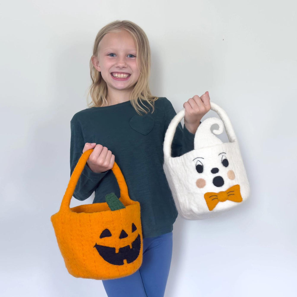 Girl holding an orange pumpkin-shaped and a white ghost-shaped Halloween bag against a plain background