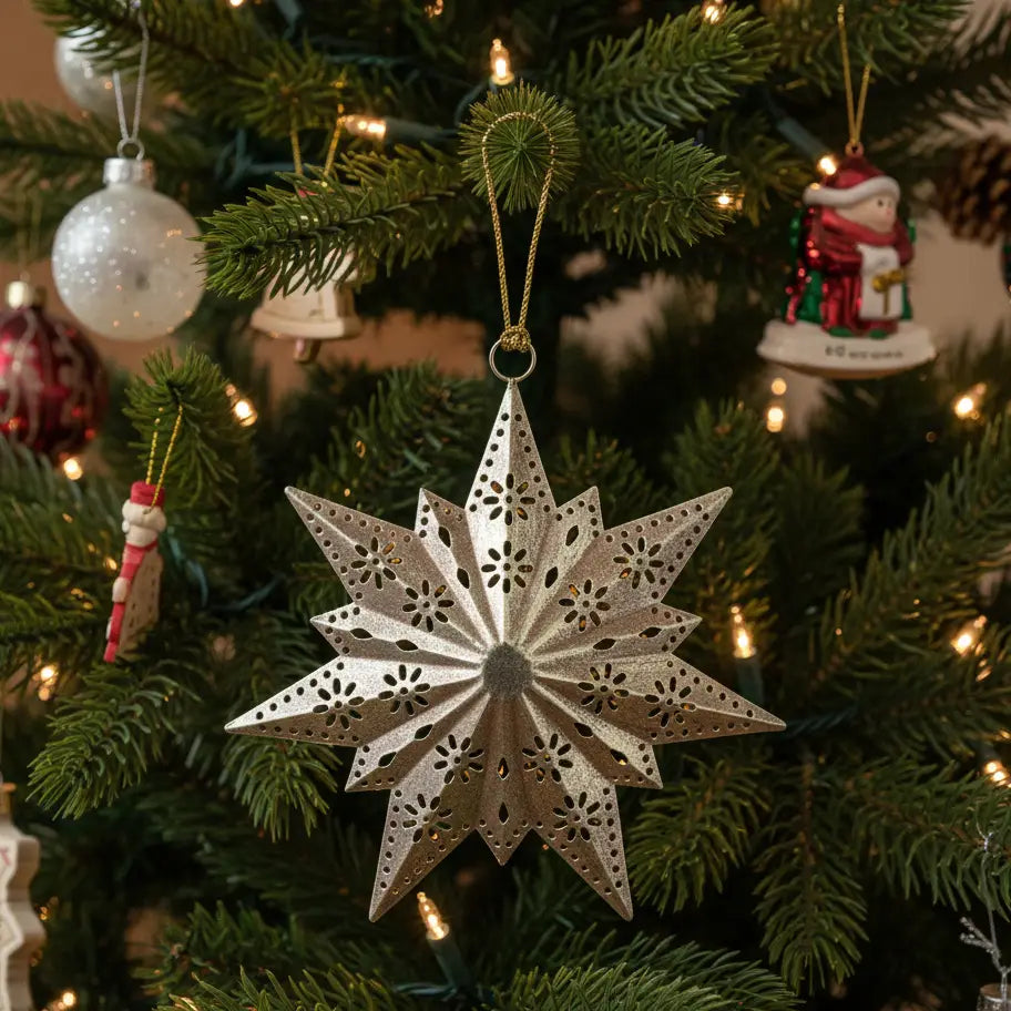 A silver metal star-shaped Christmas ornament with intricate cutout patterns hangs on a green Christmas tree with warm string lights.
