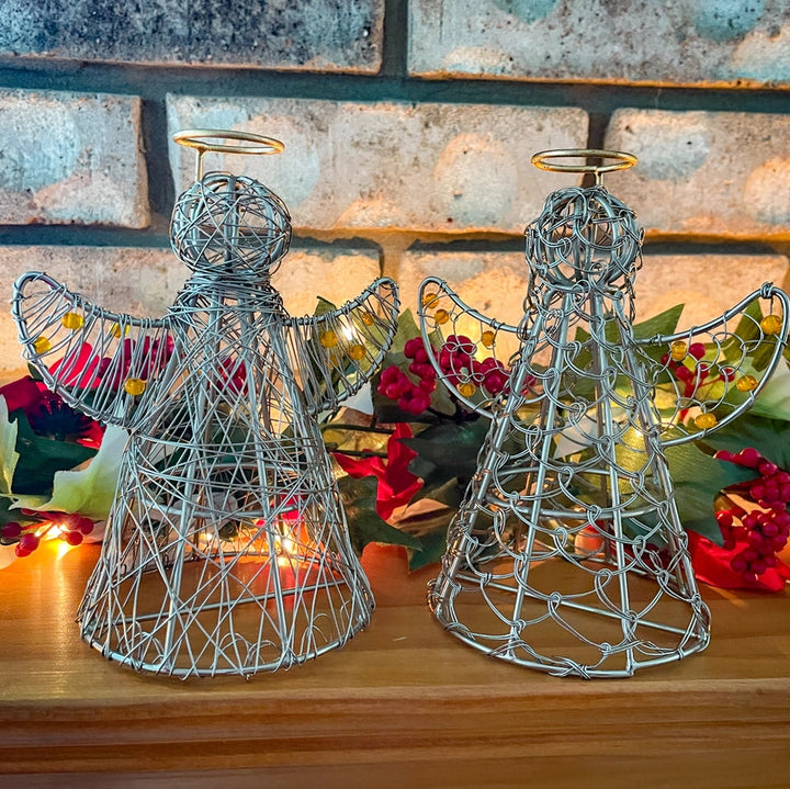 Two wire angel sculptures on a wooden surface with flowers and lights in the background.