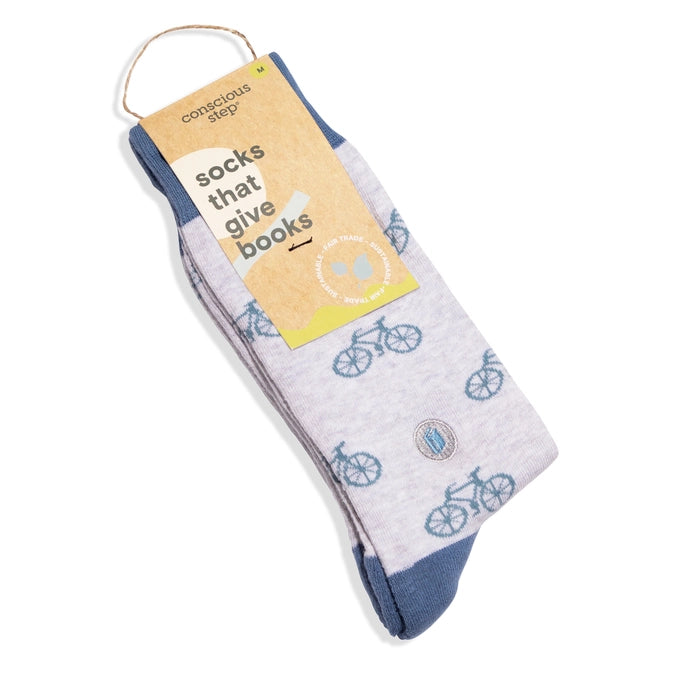 A pair of light heather gray socks with a blue bicycle pattern and navy blue cuffs, hanging from a tan paper tag that reads 'conscious step socks that give books'.