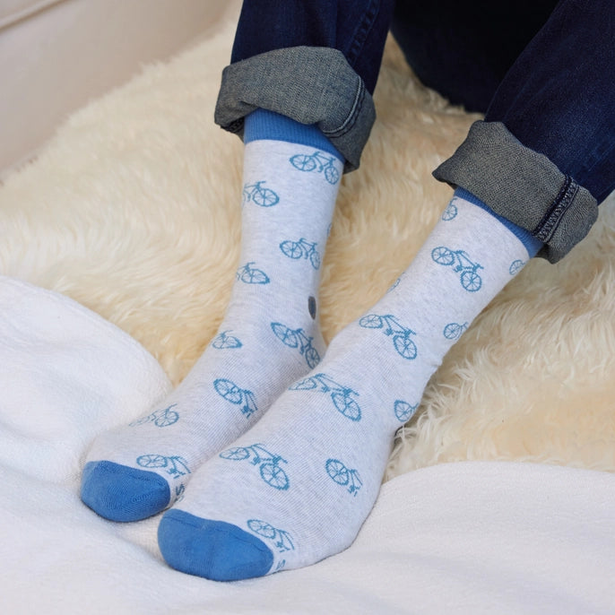 A pair of light heather gray socks with a blue bicycle pattern and navy blue cuffs, hanging from a tan paper tag that reads 'conscious step socks that give books'.
