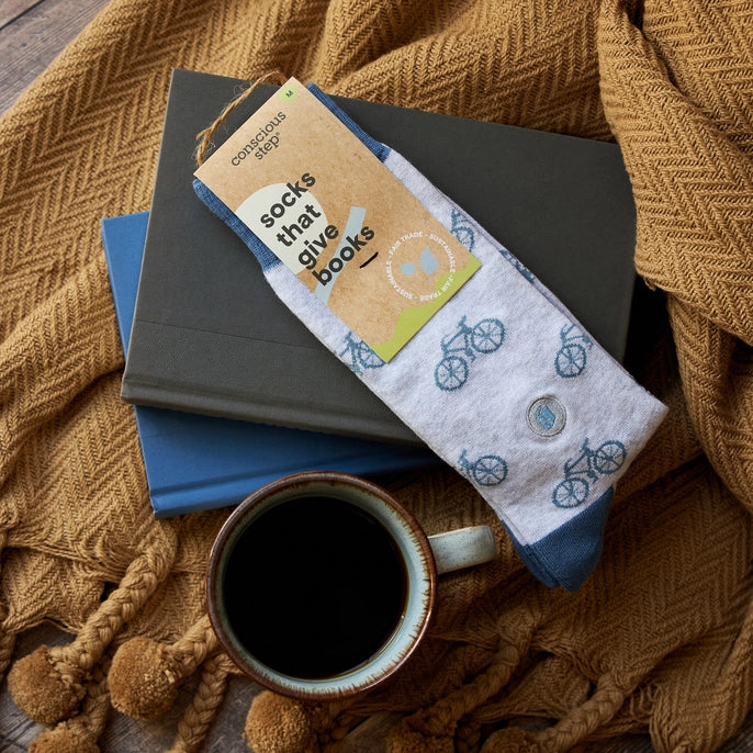 A pair of light heather gray socks with a blue bicycle pattern and navy blue cuffs, hanging from a tan paper tag that reads 'conscious step socks that give books'.
