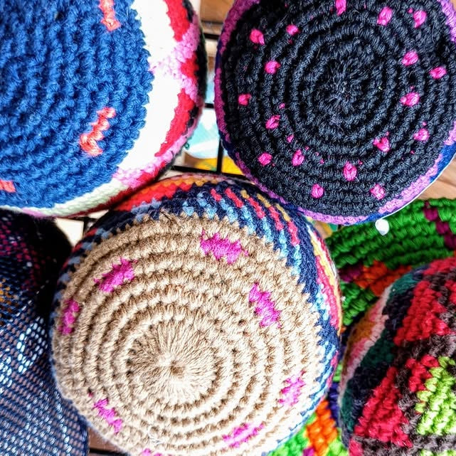 A close-up, top-down view of several colorful crocheted hacky sacks overlapping together. The visible items feature bold patterns in blue, black, pink, beige, green, and multicolor accents.