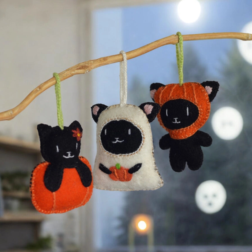 Three Halloween-themed cat ornaments hanging from a branch with a blurred background.