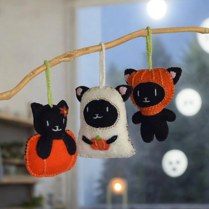 Three Halloween-themed cat ornaments hanging from a branch with a blurred background.