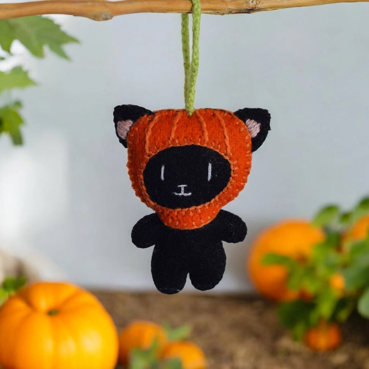 Black cat plush ornament in an orange pumpkin costume hanging from a branch with pumpkins in the background.