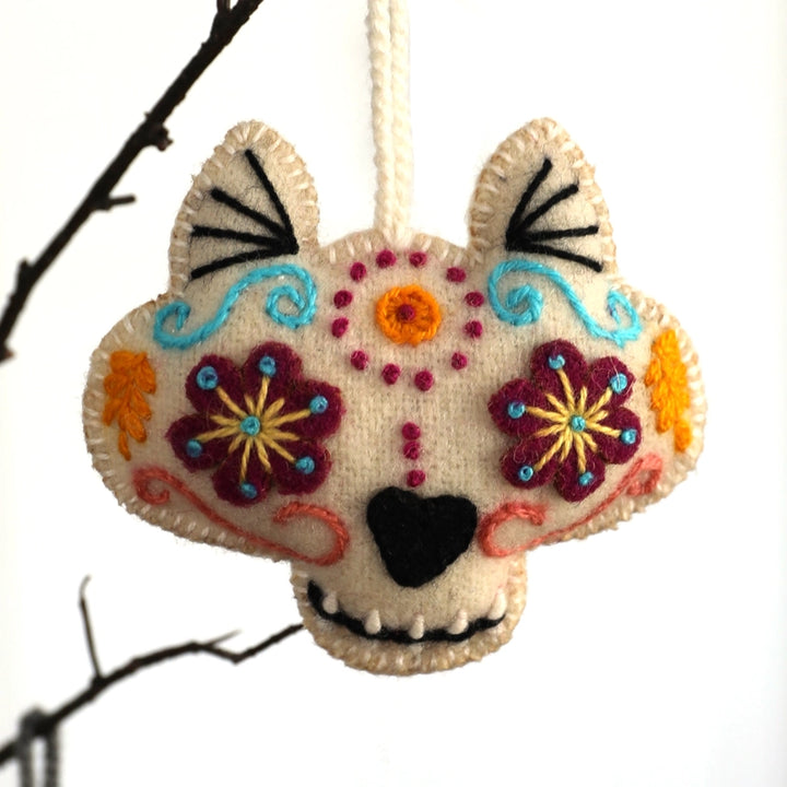 A handmade felt dog skull Christmas ornament decorated with colorful floral patterns and black embroidery, hanging from a white string against a white background with thin dark branches.