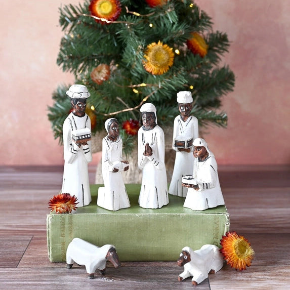 A Christmas nativity scene display featuring white figurines of three wise men and three baby Jesus figures standing on a green box, with two small white sheep at the base, set against a decorated Christmas tree and a soft pink background.