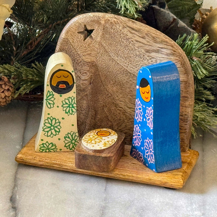 Handcrafted wooden nativity scene display featuring a carved wooden stable backdrop, a cream-colored baby cradle with green floral patterns, a bright blue baby cradle with white floral patterns, and a small round wooden box, all arranged on a light wood base with evergreen branches in the background.