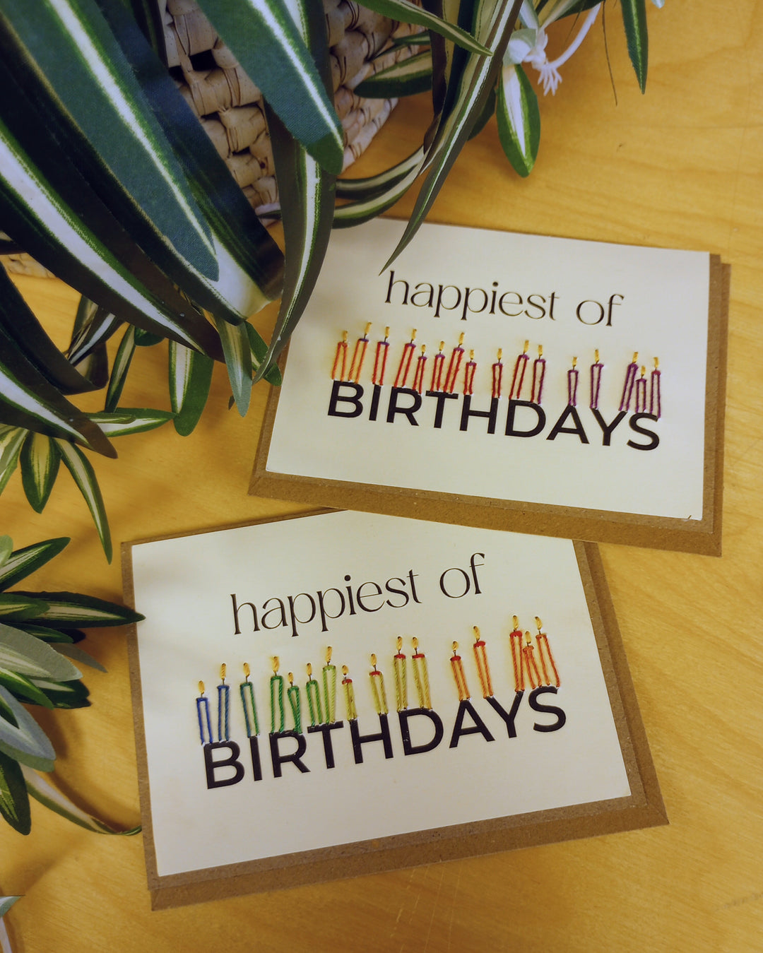 Two 'happiest of birthdays' cards with candles on a wooden surface with plants.