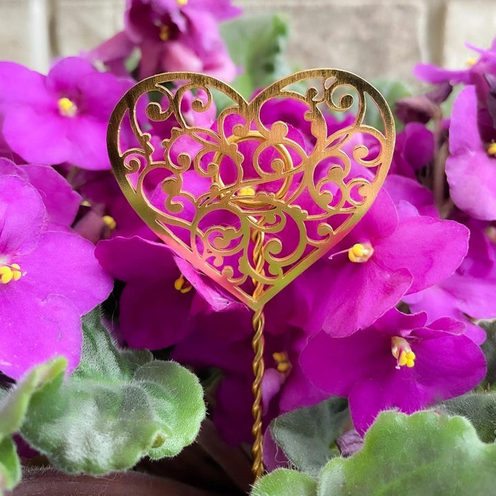 Decorative gold heart-shaped plant stake among purple African violets with a blurred brick wall background.