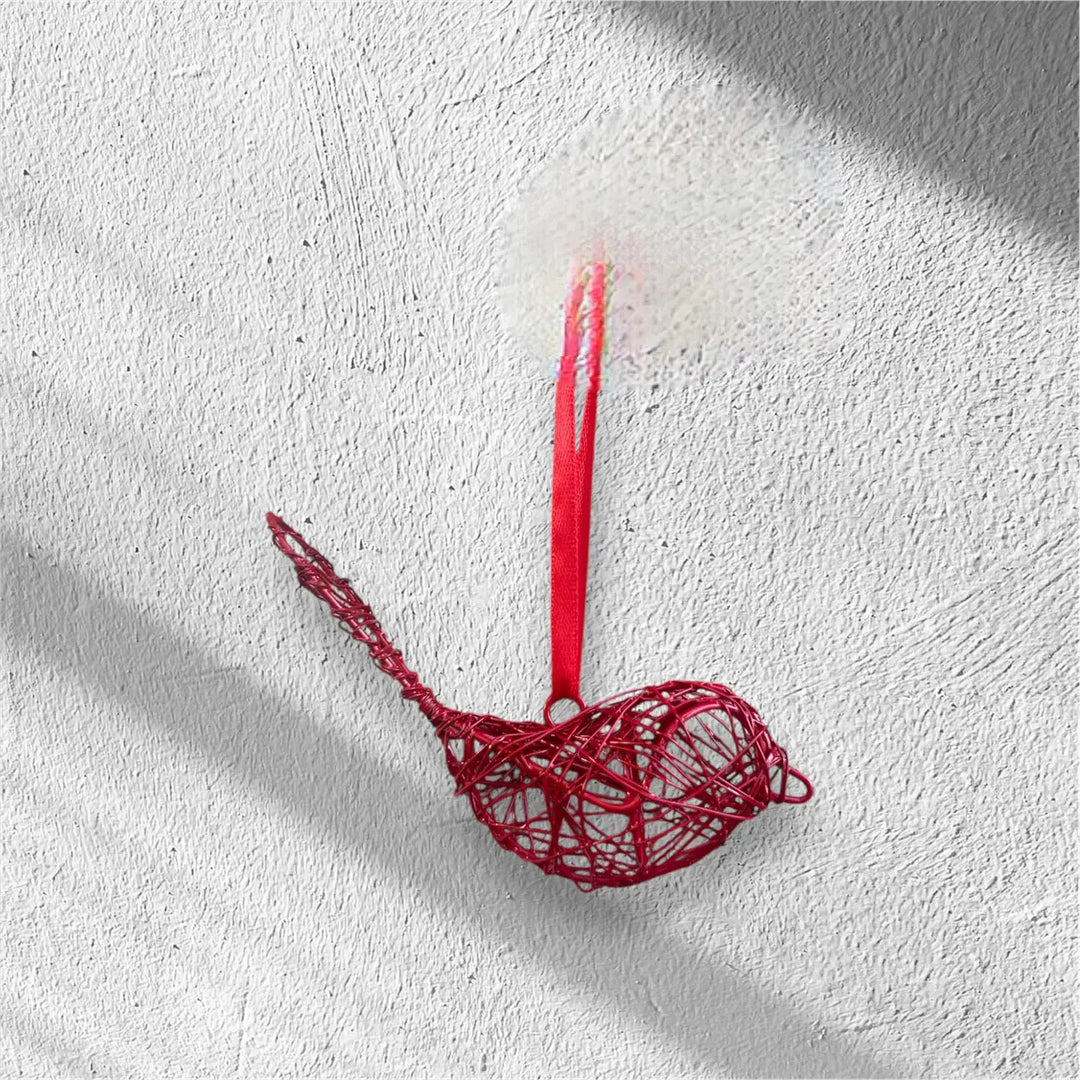 A red wire bird ornament hanging from a red ribbon against a textured white wall with soft shadows.