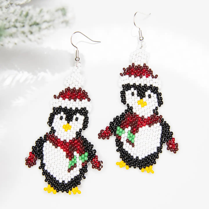 A pair of handmade beaded dangle earrings shaped like penguins, decorated with black and white beads, red scarves and hats, yellow beaks and feet, and small green accents, shown against a white background with a soft blurred holiday branch.
