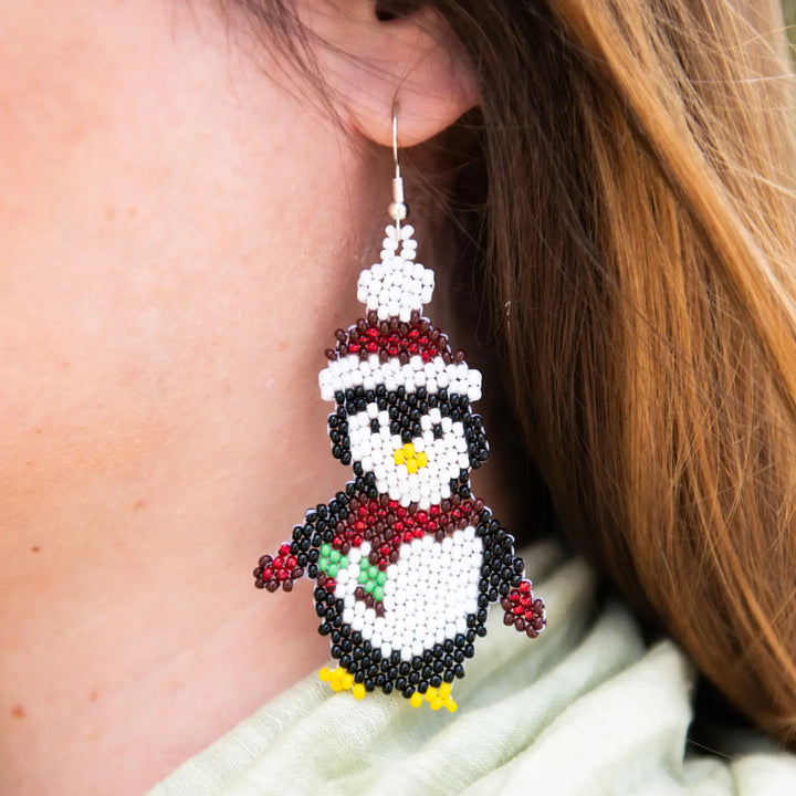 A close-up of a person's ear wearing a beaded penguin earring. The earring is made from white, black, and red seed beads with small yellow accents for the beak and feet, and a tiny green accent for the chest detail.