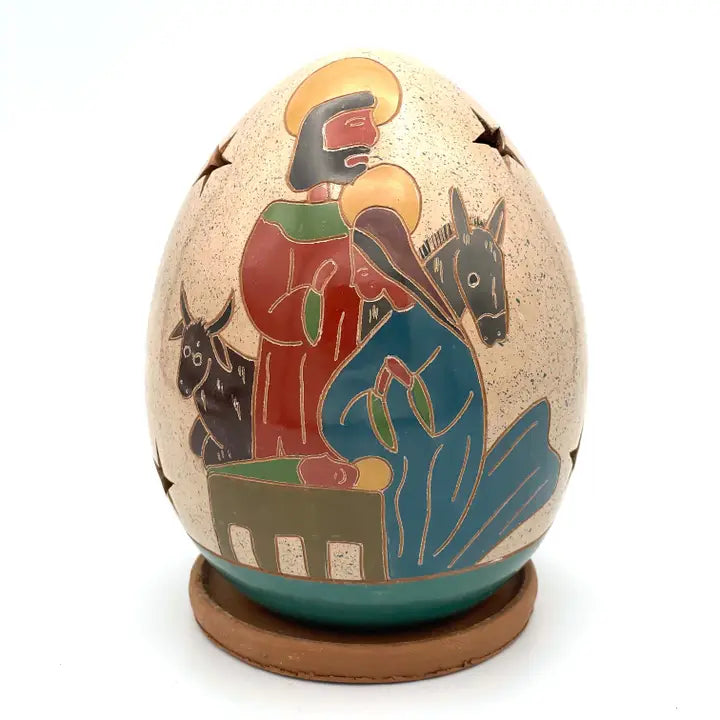 A decorative nativity egg with hand-painted figures of Mary, Joseph, baby Jesus, and animals, displayed on a small round wooden base against a white background.