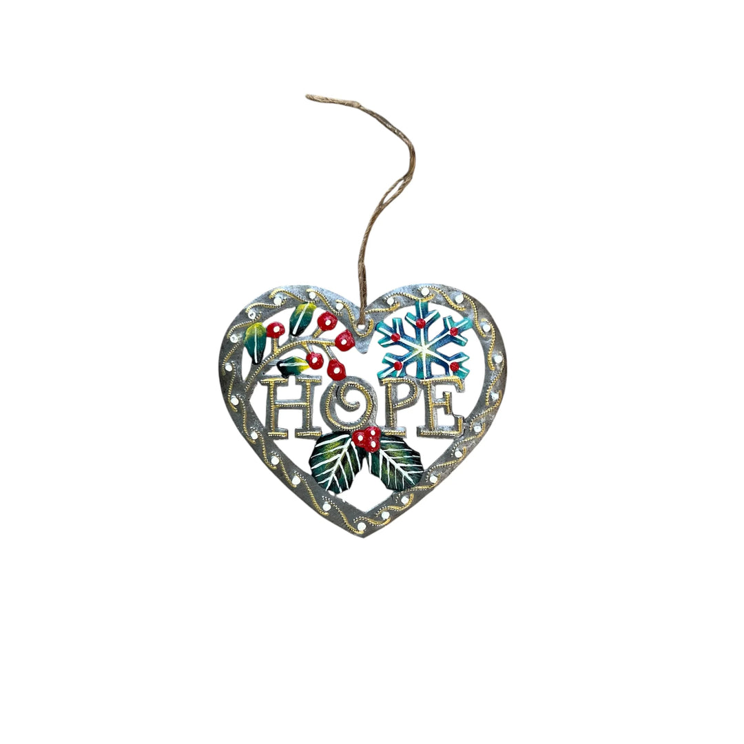 A heart-shaped metal Christmas ornament with the word 'HOPE' in the center, decorated with a snowflake, holly leaves, berries, and a hanging twine string, shown on a plain white background.