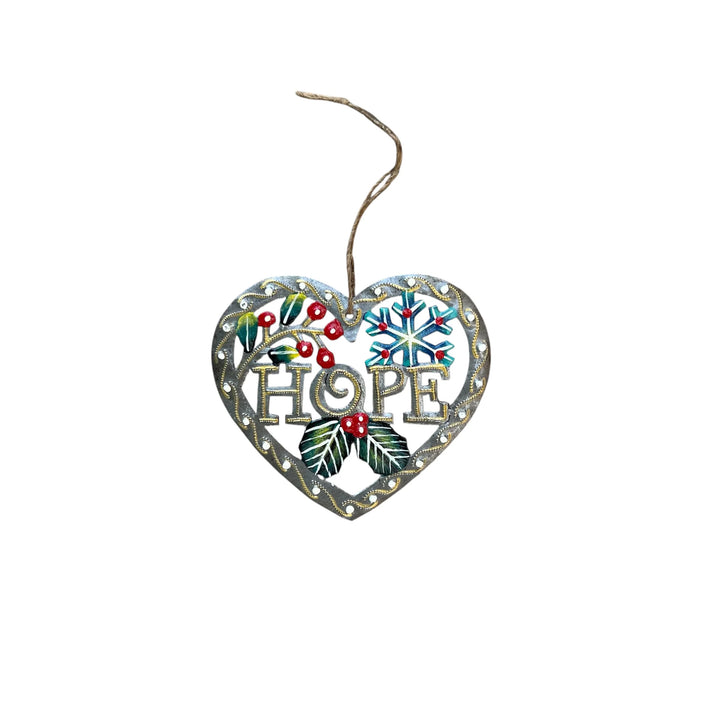 A heart-shaped metal Christmas ornament with the word 'HOPE' in the center, decorated with a snowflake, holly leaves, berries, and a hanging twine string, shown on a plain white background.