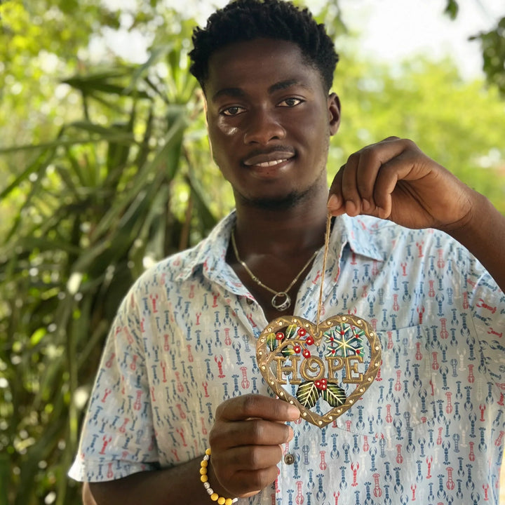 A smiling young man outdoors holds up a heart-shaped gold pendant necklace with the word 'HOPE' in gold letters and colorful red, green, and blue details. He is wearing a light blue patterned short-sleeve button-up shirt and a gold chain necklace.