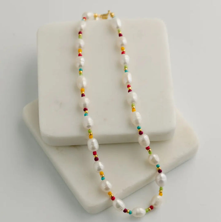 A beaded necklace with white fresh water pearls and small multicolored beads, displayed on a white marble block against a light gray background.