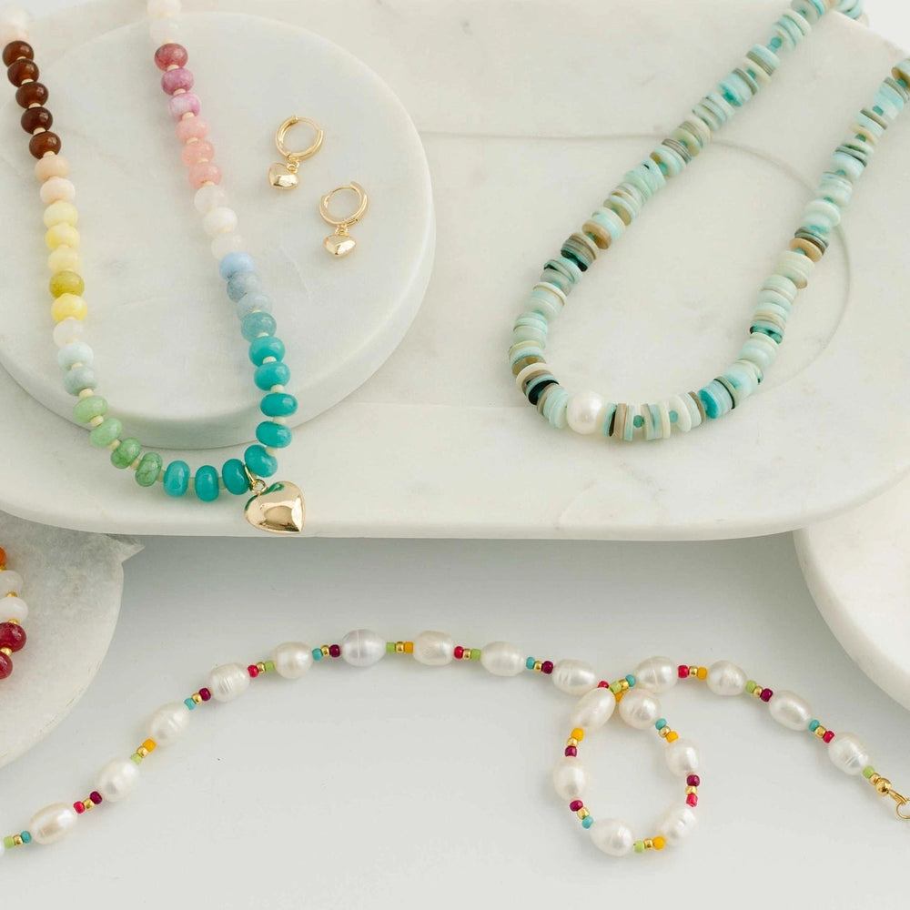 A styled flat-lay of multiple beaded jewelry pieces on white marble trays, including necklaces, bracelets, and gold-tone earrings, with colorful beads and turquoise accents on a white background.