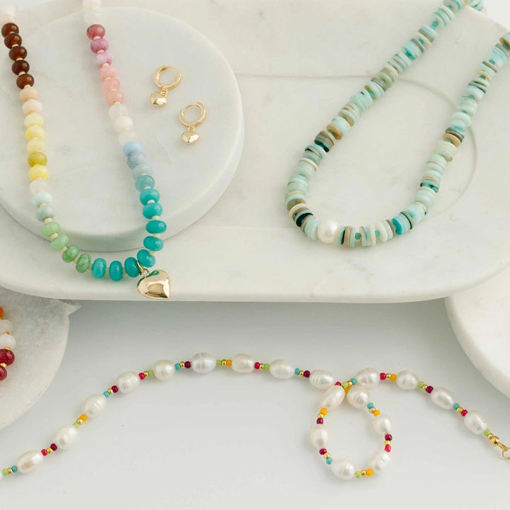A styled flat-lay of multiple beaded jewelry pieces on white marble trays, including necklaces, bracelets, and gold-tone earrings, with colorful beads and turquoise accents on a white background.