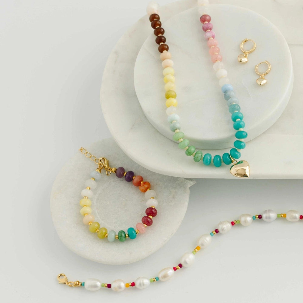 A styled flat-lay of multiple beaded jewelry pieces on white marble trays, including necklaces, bracelets, and gold-tone earrings, with colorful beads and turquoise accents on a white background.