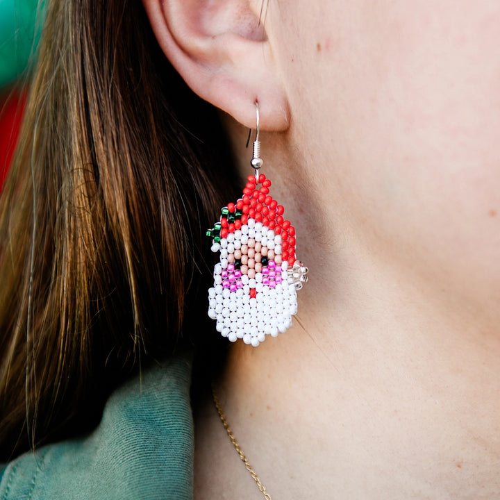A close-up of a person wearing a beaded Santa Claus earring. The earring features white seed beads for the beard, red beads for the hat, and small pink, green, and black beads for the face details, hanging from a silver fishhook ear wire.