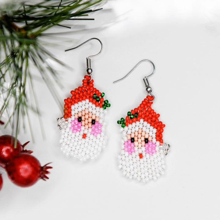 A pair of beaded Santa Claus face drop earrings with silver fishhook wires, displayed on a clean white background with pine needles and red ornaments as decorative props.