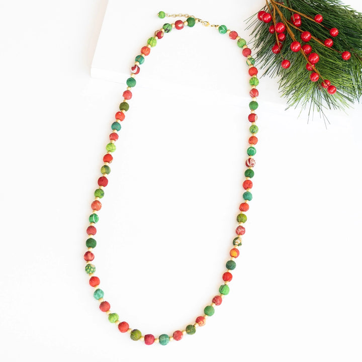 A long beaded necklace made of round beads in red, and green, arranged in a loop on a white background with a pine branch and red berries in the top right corner.