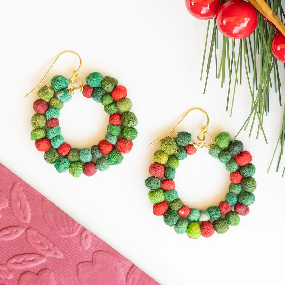 A pair of handmade hoop earrings made from textured green and red kantha beads, attached to gold fishhook wires, displayed on a white background with Christmas-themed flat-lay props.