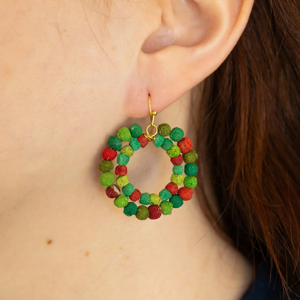 A close-up of a person's ear wearing a handmade circular dangle earring made of small textured green and red beads with a gold-tone hook.