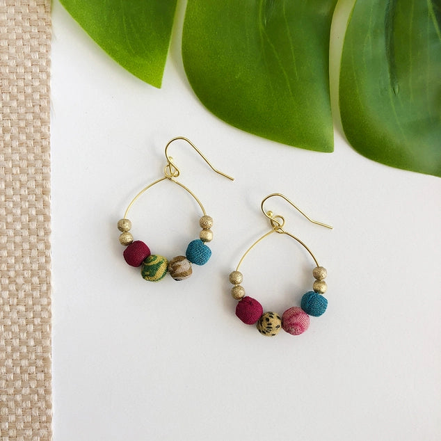 Kantha gold hoop earrings with colorful beads on a white surface with green leaves in the background