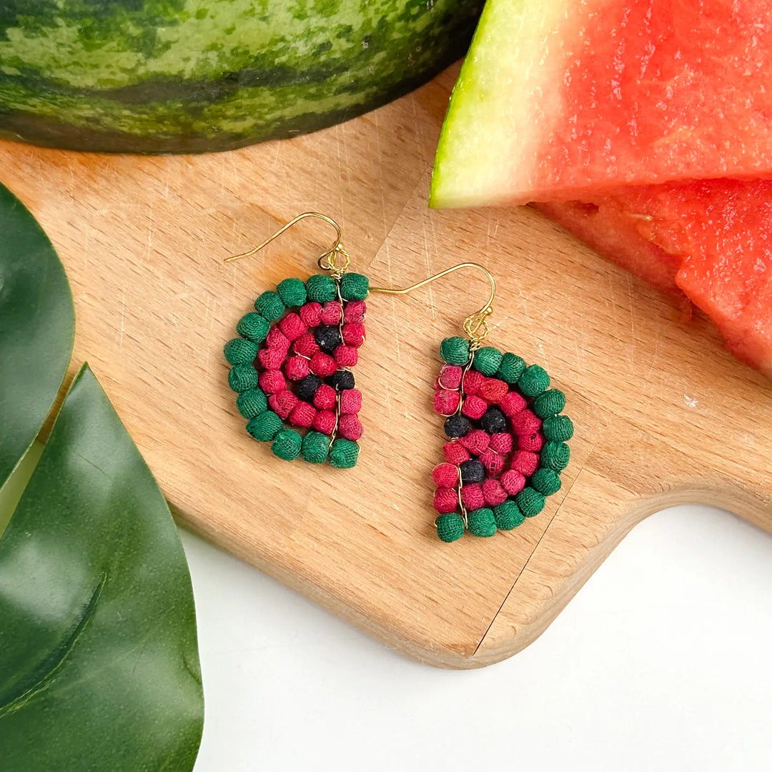 Katha watermelon-themed earrings on a wooden cutting board with watermelon slices and leaves.