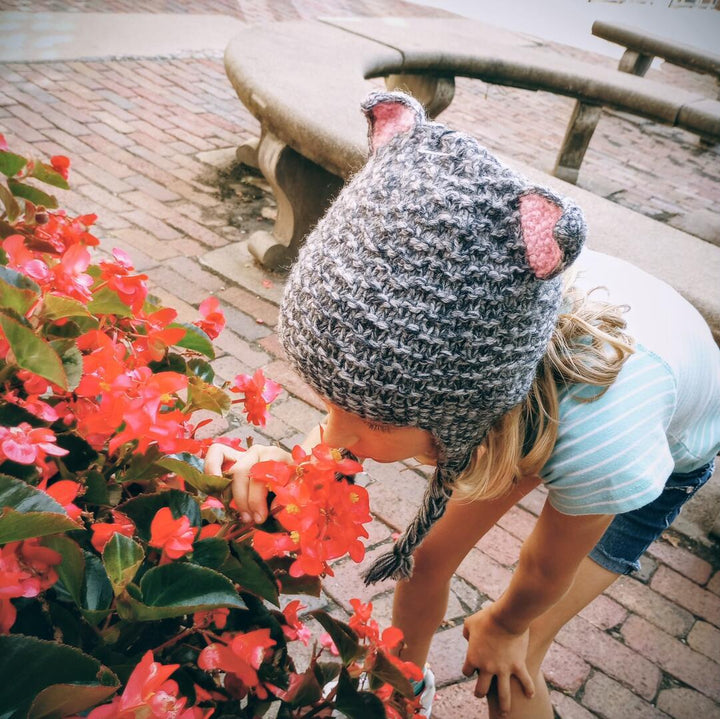Child wearing a knitted hat with animal ears, smelling flowers outdoors.
