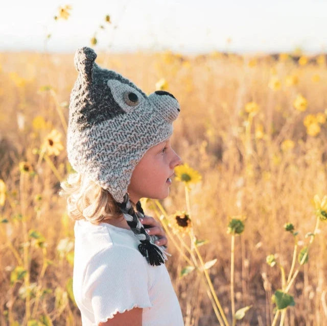 A child in profile wearing a crocheted wolf-style hat with ear-like protrusions and a braided strap, standing in a sunflower field.