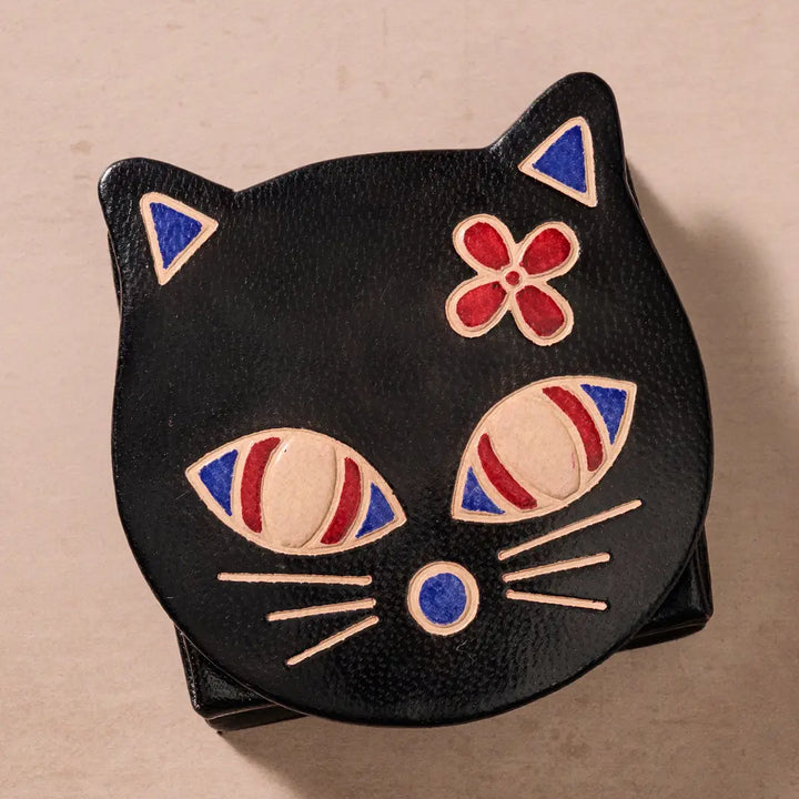 A black faux-leather cat-shaped coin purse with hand-painted red, blue, and beige details on its face, photographed on a neutral beige background.