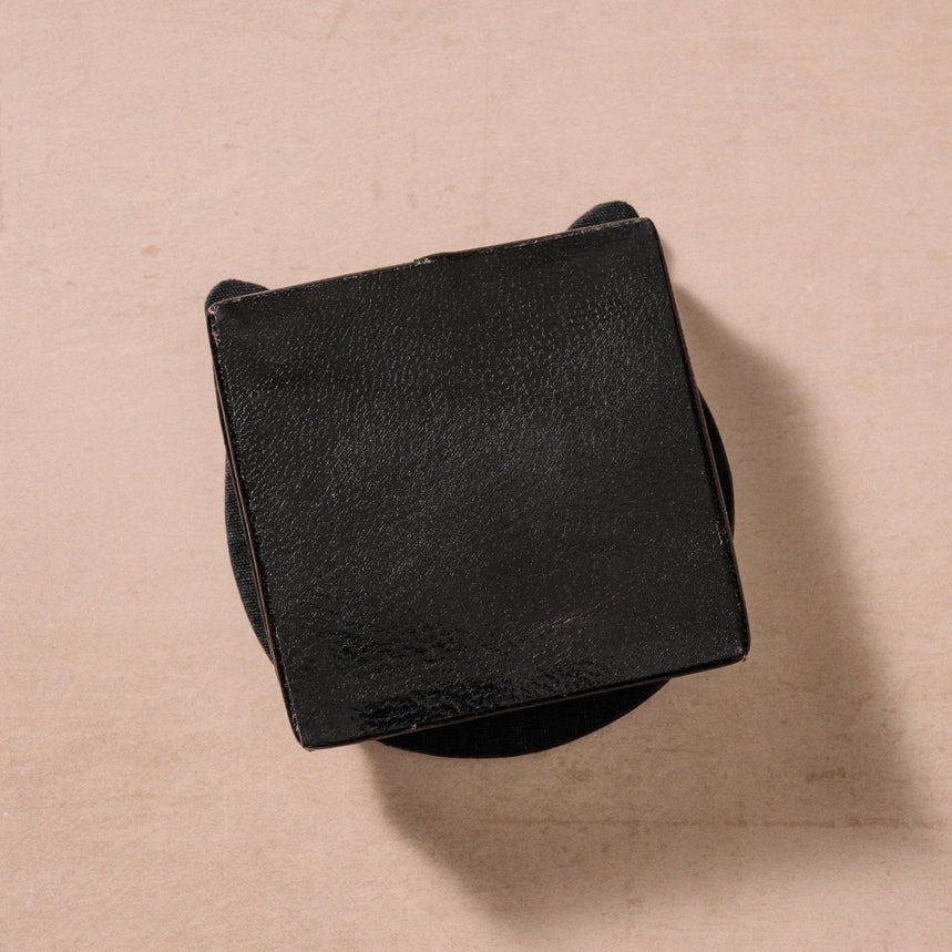 A black textured square box with rounded corners, photographed from above on a warm beige background.