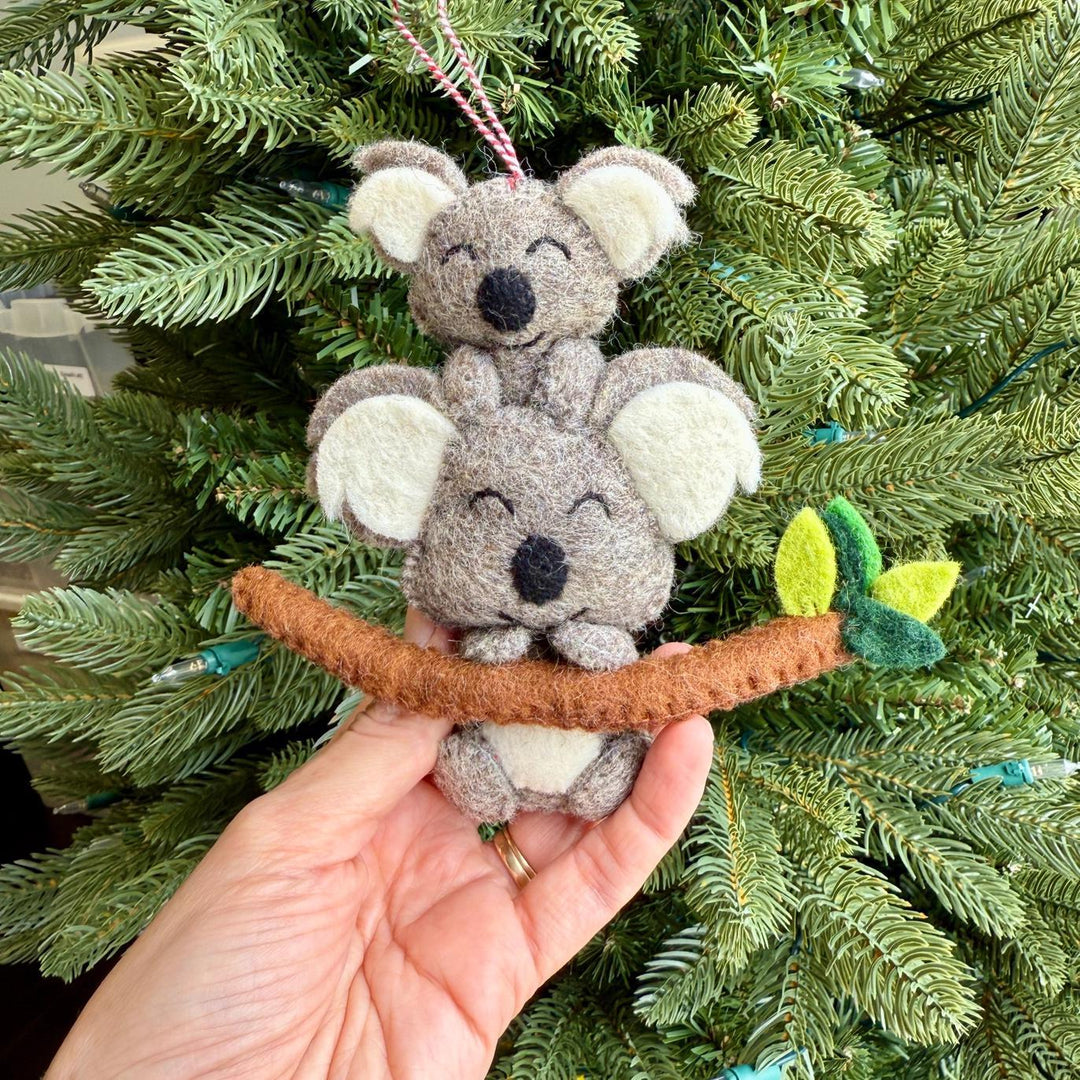 Two small felt koala ornaments on a branch held in front of a Christmas tree.