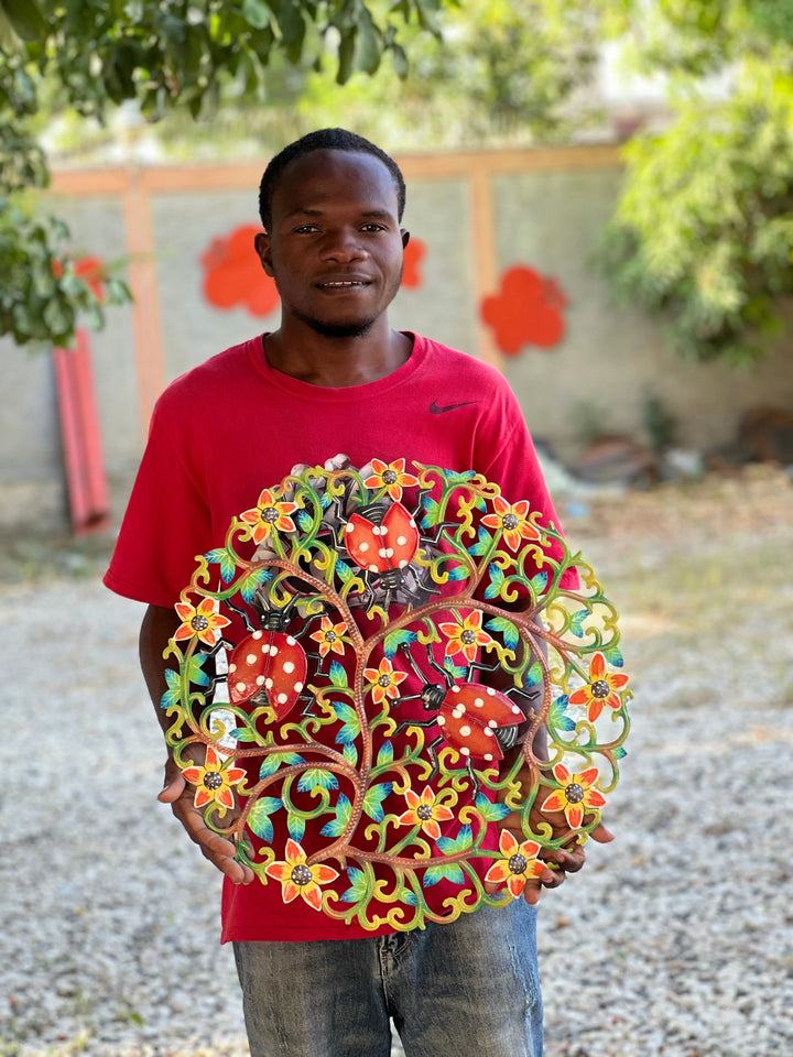 Person holding a colorful metal art piece outdoors with flowers and ladybugs in the background
