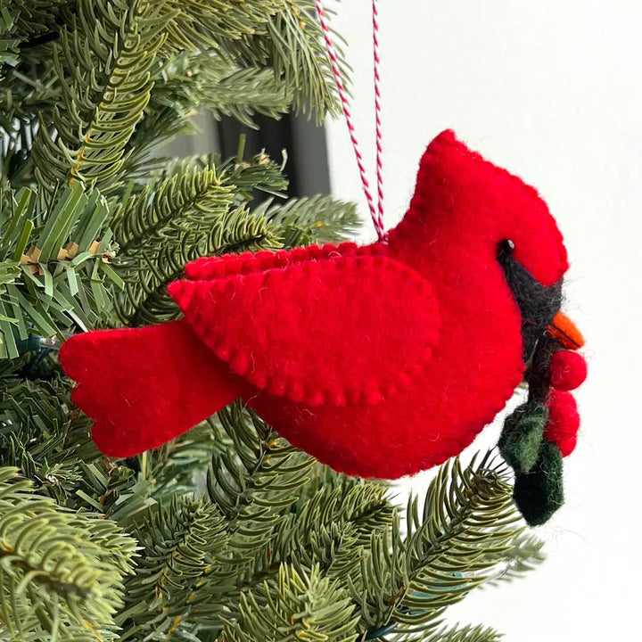 A red felt cardinal Christmas ornament hanging from a red-and-white string, positioned among green pine tree branches with a white background visible behind it.