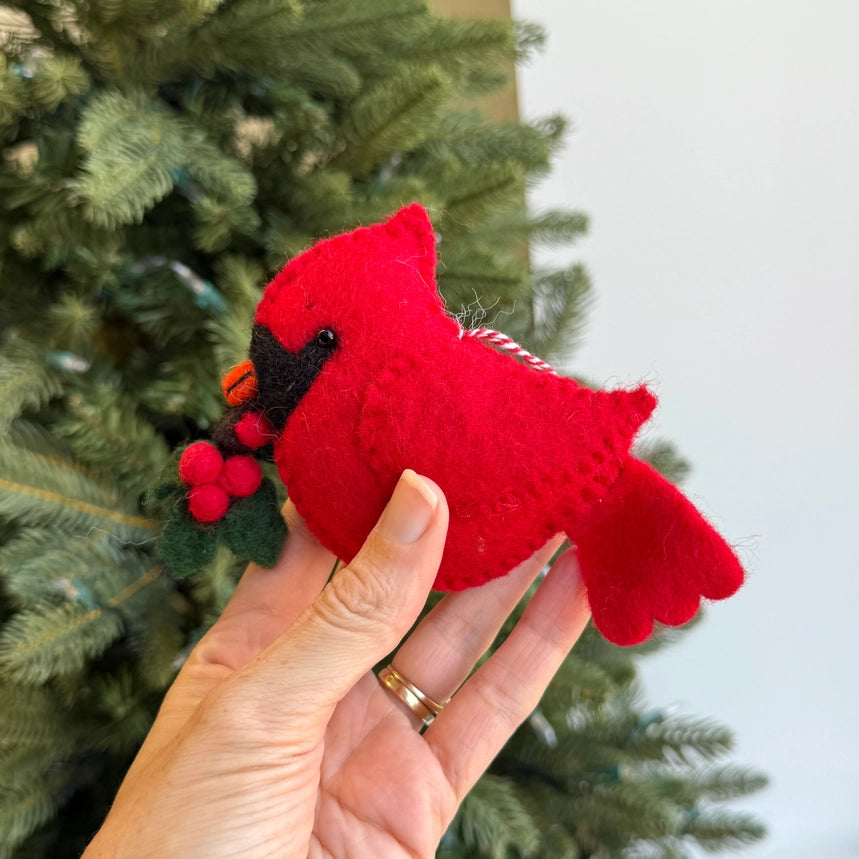 A hand holds a red felt cardinal ornament with a black beak and a small holly sprig, with a Christmas tree and light wall in the background.
