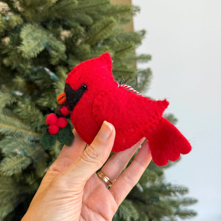A hand holds a red felt cardinal ornament with a black beak and a small holly sprig, with a Christmas tree and light wall in the background.