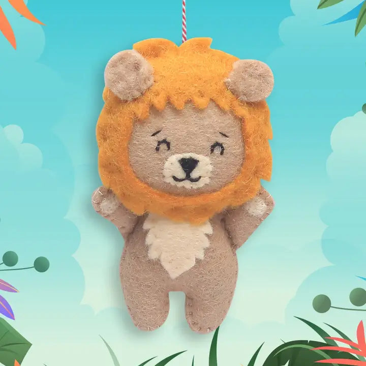 A plush lion hanging ornament made of felt fabric, featuring a golden-orange mane, beige body, and embroidered facial details, suspended against a colorful illustrated background.